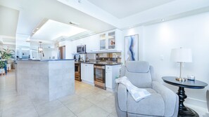 A modern kitchen with white cabinetry, a central island, and a dining area with a grey armchair and a small table. - #7306 Towers 8&9 Oceanfront 2/2 condo (Ponce Inlet)