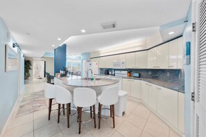 A modern kitchen with a central island, white bar stools, and a view into the adjacent living area. - Towers at Ponce Inlet 1206 (Ponce Inlet)