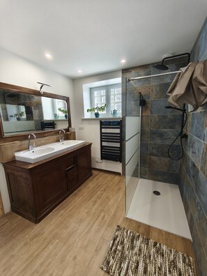 Un baño moderno con tocador doble lavamanos, cabina de ducha con mampara de vidrio y ventana que proporciona luz natural.