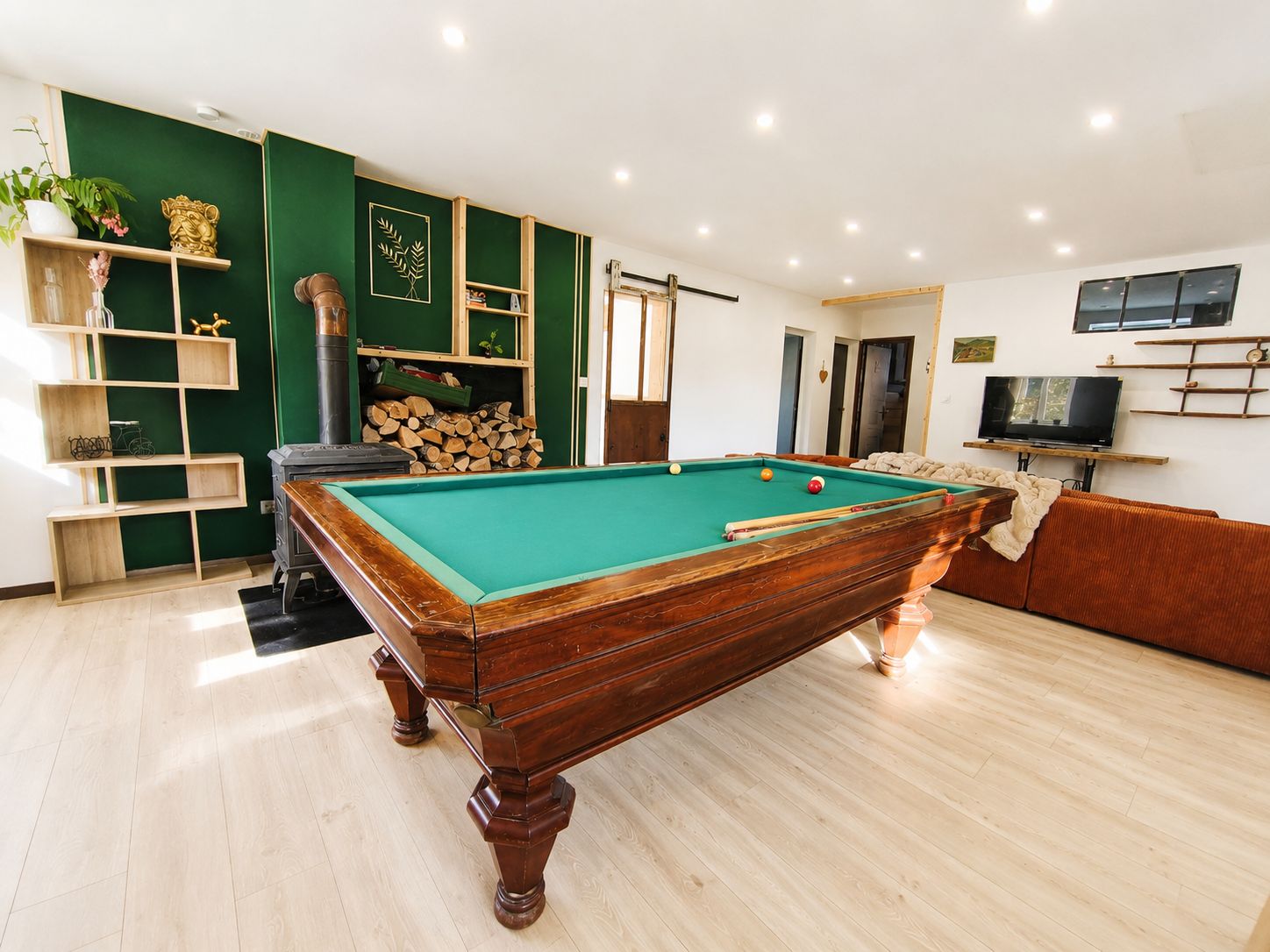 A pool table in a room with a green accent wall, wooden shelves, and a television mounted on the wall.