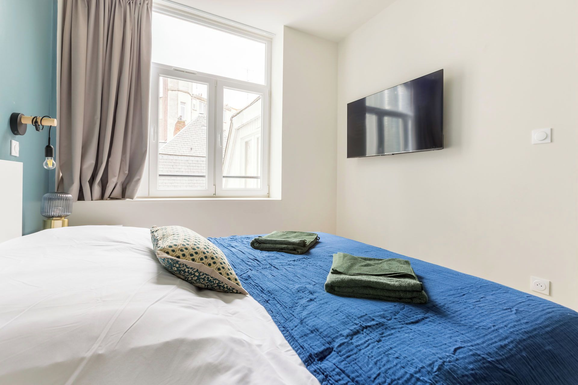 Une chambre d’hôtel avec un lit, un téléviseur fixé au mur, une fenêtre avec des rideaux et une vue sur les bâtiments environnants.