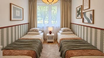 Ein Hotelzimmer mit zwei Betten, einem Fenster mit Vorhängen, einem Nachttisch mit Lampe sowie gerahmten Kunstwerken an den Wänden.