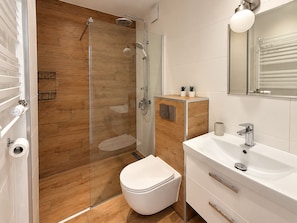 A modern bathroom with a glass shower enclosure, a toilet, a sink with a mirror, and a small cabinet with plants. - Family Apartment with Balcony (Gdańsk)
