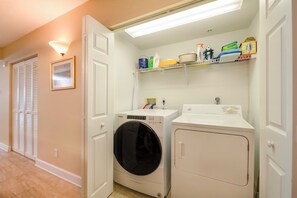 A laundry room with a front-loading washer and dryer, a shelf with cleaning supplies, and a door with a framed picture. - #6410 Towers Oceanview 2/2 Condo (Ponce Inlet)