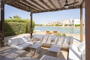 Ein überdachter Außenbereich mit weißen Sofas und Tischen mit Blick auf einen Strand und Gebäude.