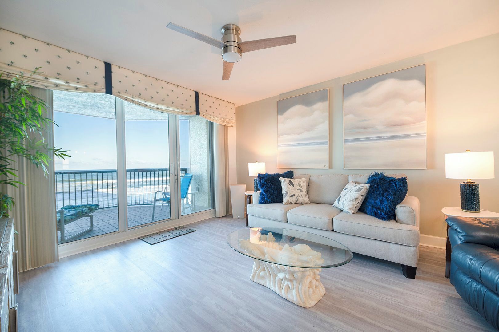 A living room with a glass coffee table, a sofa with blue and white cushions, a ceiling fan, and a view of the beach through a sliding glass door.