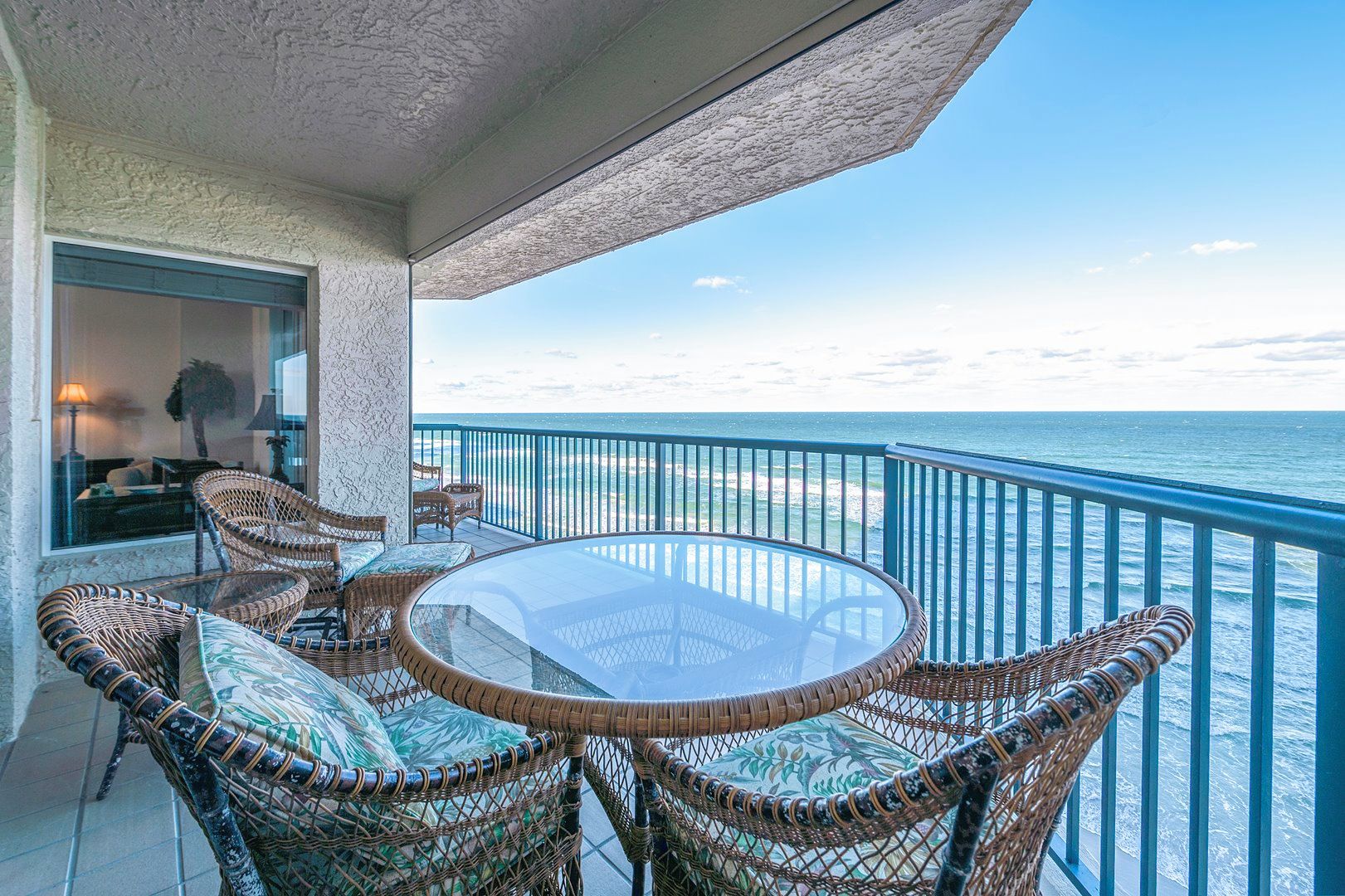 A balcony with wicker furniture and a glass table overlooking the sea.