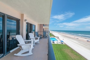 Ein Balkon mit weißen Schaukelstühlen, ein Poolbereich mit Sonnenschirmen und Blick auf den Strand.