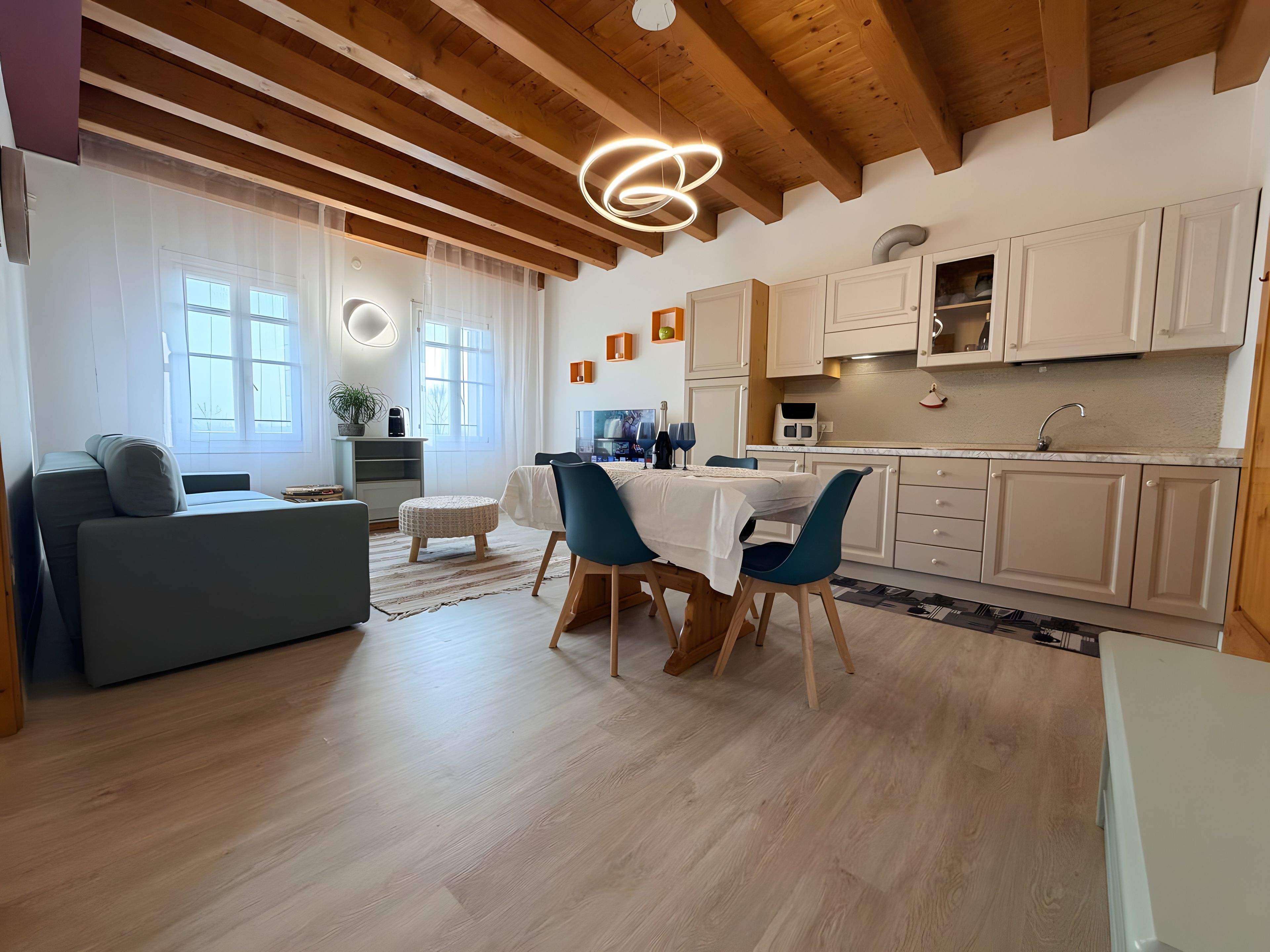 A modern living room with a dining area, wooden ceiling beams, and a kitchen with white cabinets.