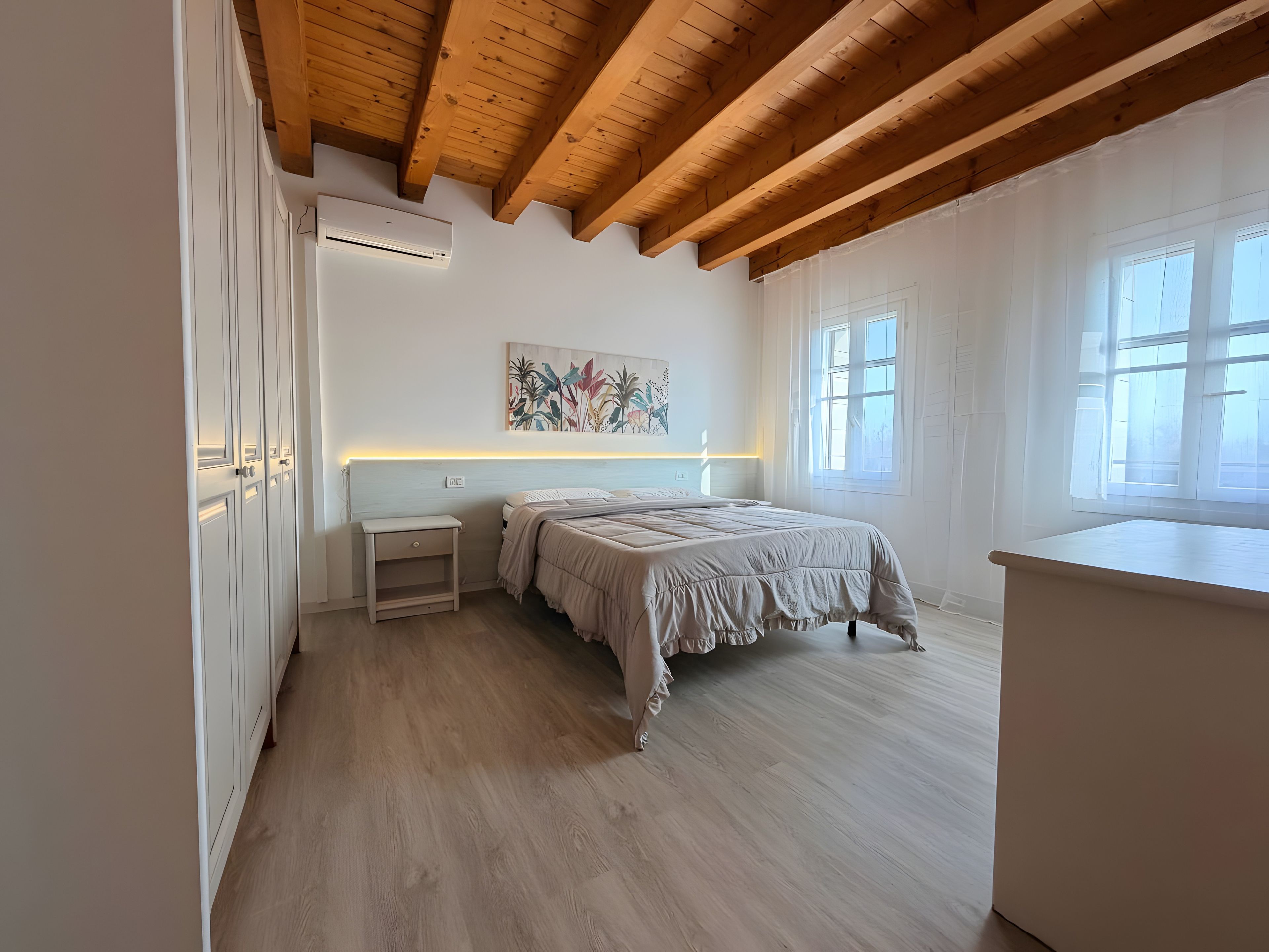 Une chambre avec un plafond en bois, un lit doté d’une tête de lit blanche, une table de chevet et une fenêtre.