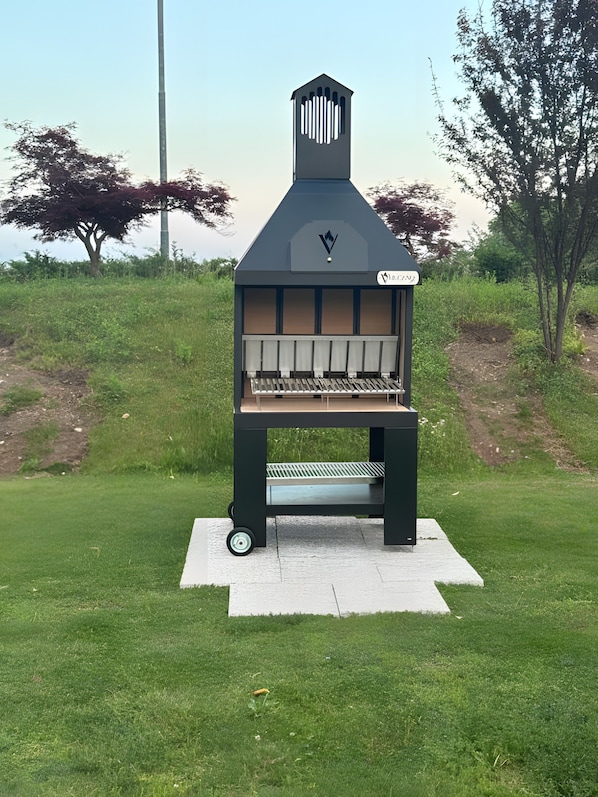 A portable barbecue grill with a chimney, wheels, and a label in a grassy outdoor area. - Apartment 'Alloggio 2 - Monolocale' with shared pool, Wi-Fi and air conditioning (Romano d'Ezzelino)