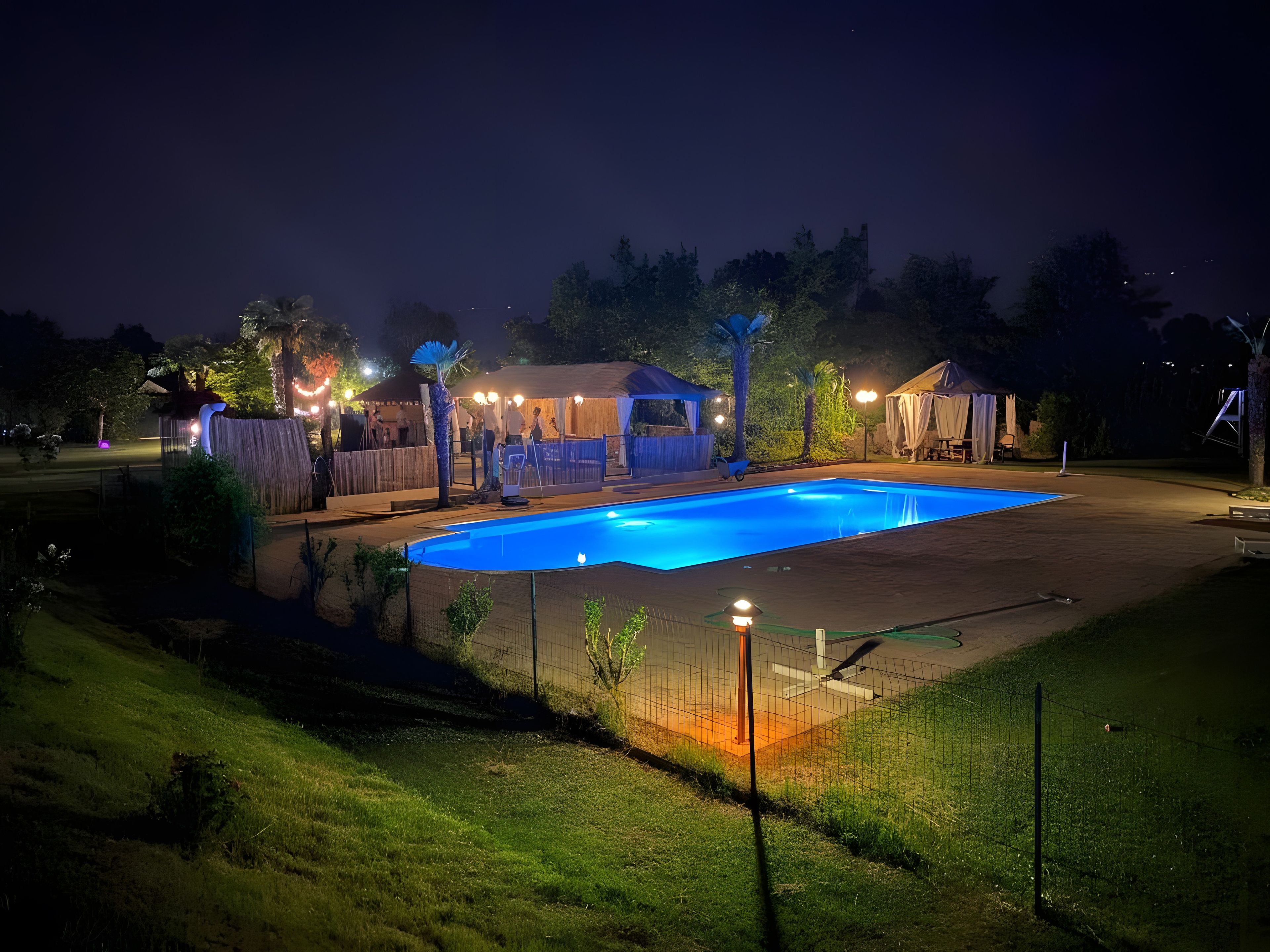 A swimming pool with a fence, surrounded by a grassy area and a covered seating area.