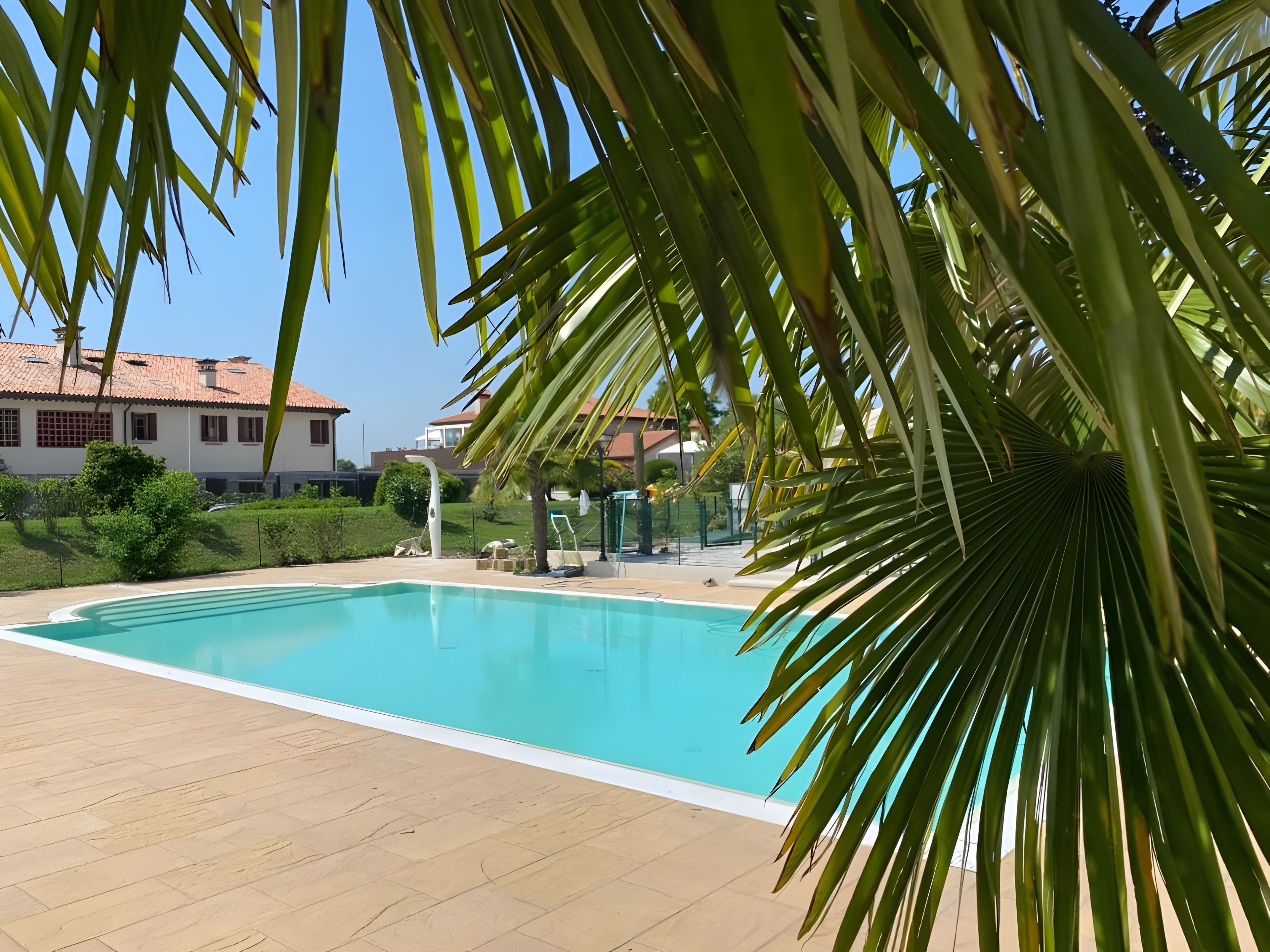 A swimming pool with a tiled deck, surrounded by greenery and a clear sky.