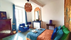 Un dormitorio con una cama, un sillón azul, un aparador, un espejo y una ventana.