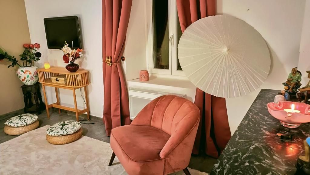 Una habitación con una silla rosa, una mesa redonda con una vela, un televisor montado en la pared y una ventana con cortinas.