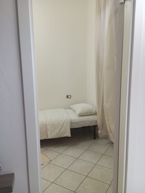 Ein kleiner, schmaler Raum mit einem Einzelbett, einem Vorhang und einem Fliesenboden.