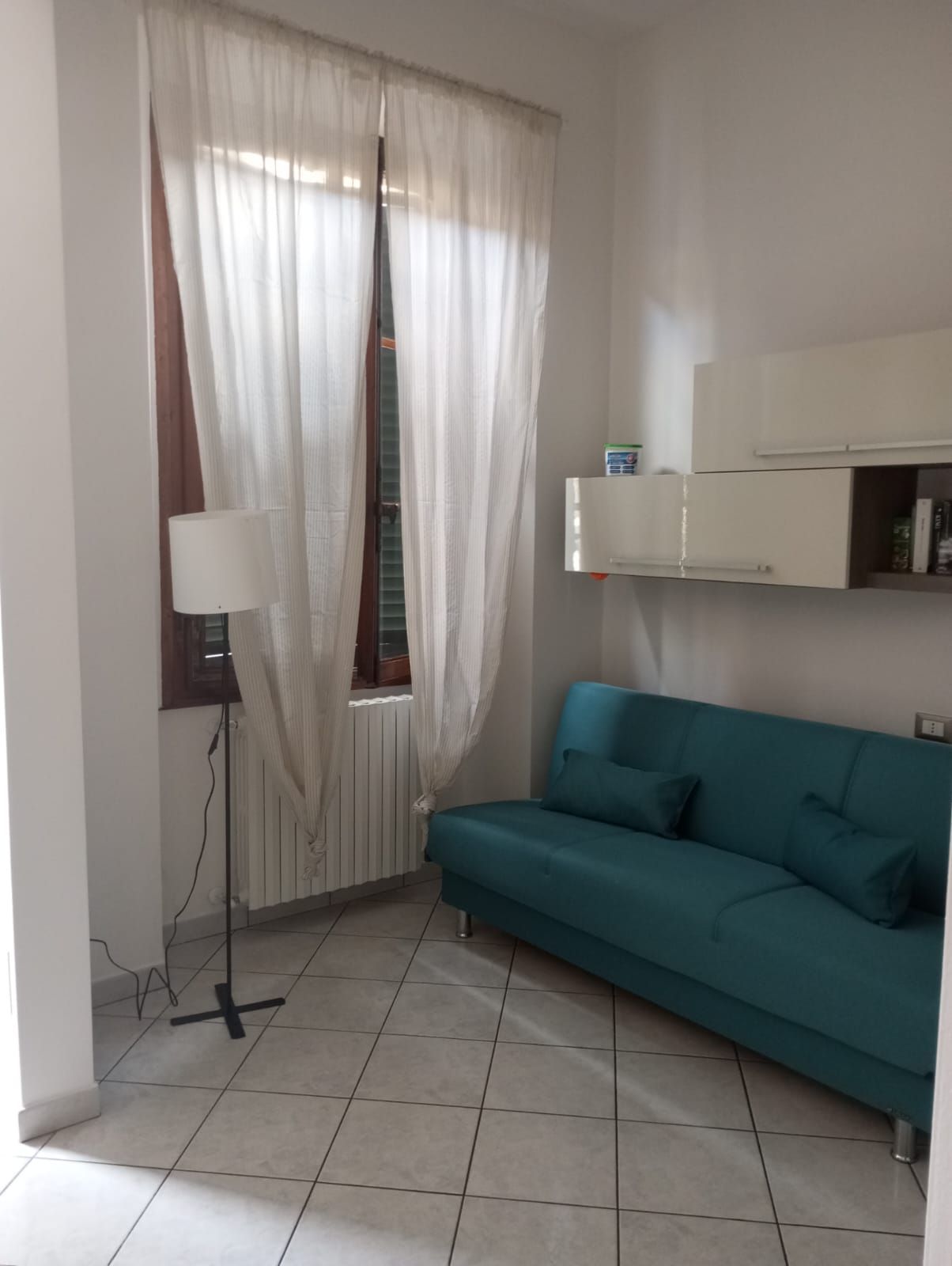 Ein Zimmer mit einem türkisfarbenen Sofa, einem weißen Heizkörper, einer weißen Lampe und einem weißen Schrank mit einem kleinen Fernseher.