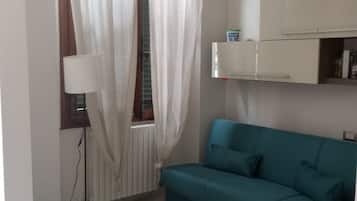 Una habitación con un sofá color turquesa, un radiador blanco, una lámpara blanca y un mueble blanco con una pequeña televisión.