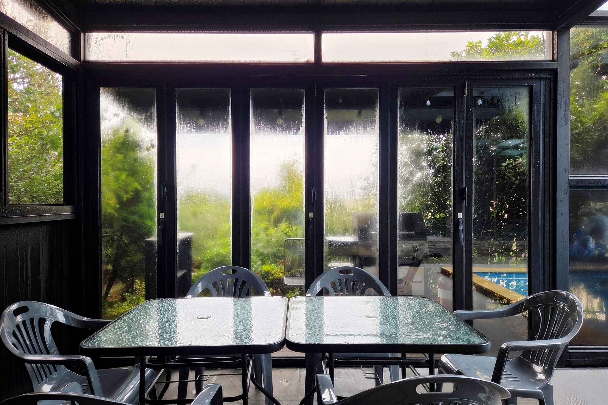 Un patio cubierto con cerramiento de vidrio, que incluye una mesa y sillas, y vistas al verdor exterior.