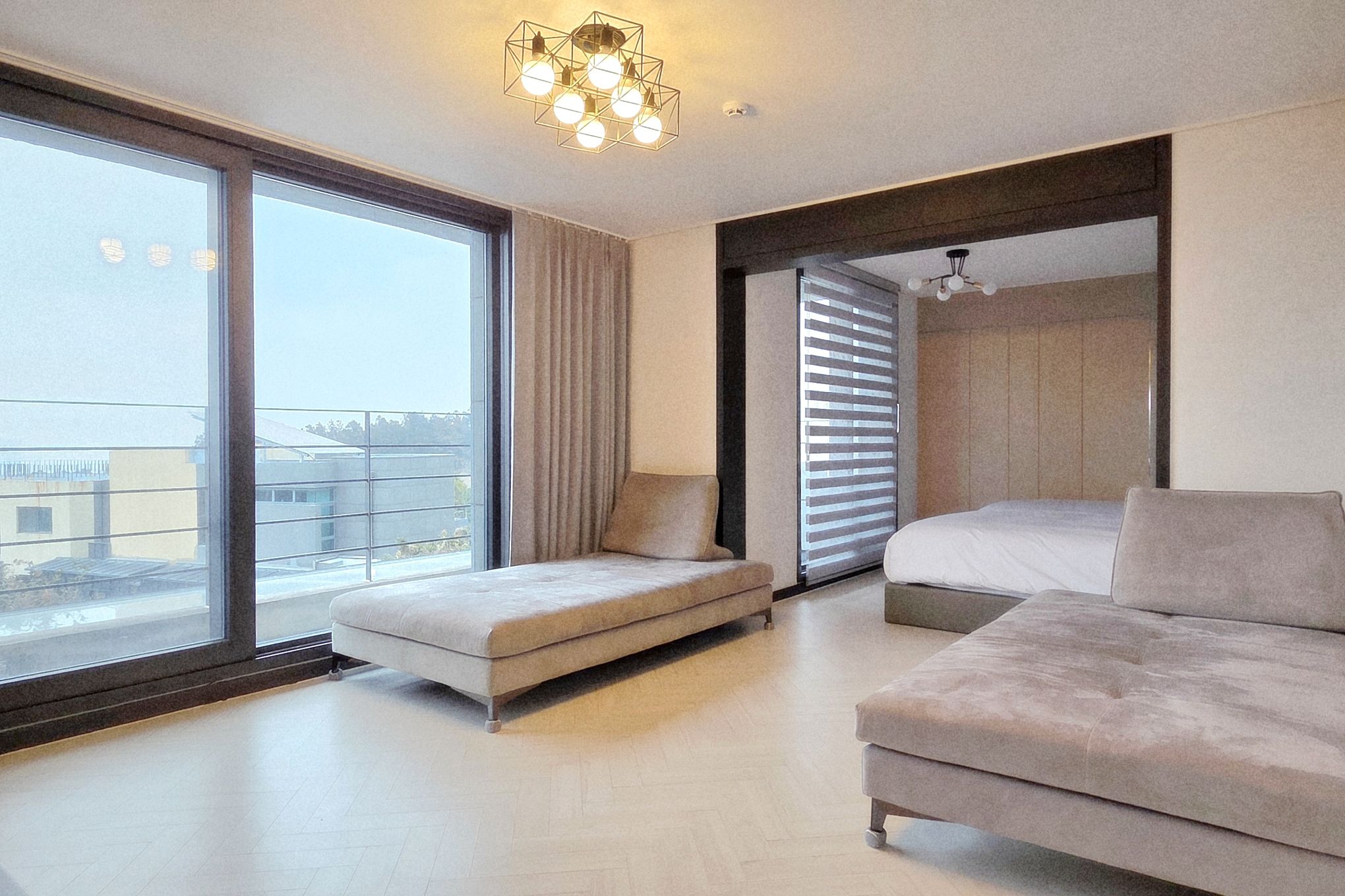 A modern hotel room with a large sliding glass door leading to a balcony, a sofa, a bed, and a ceiling light fixture.