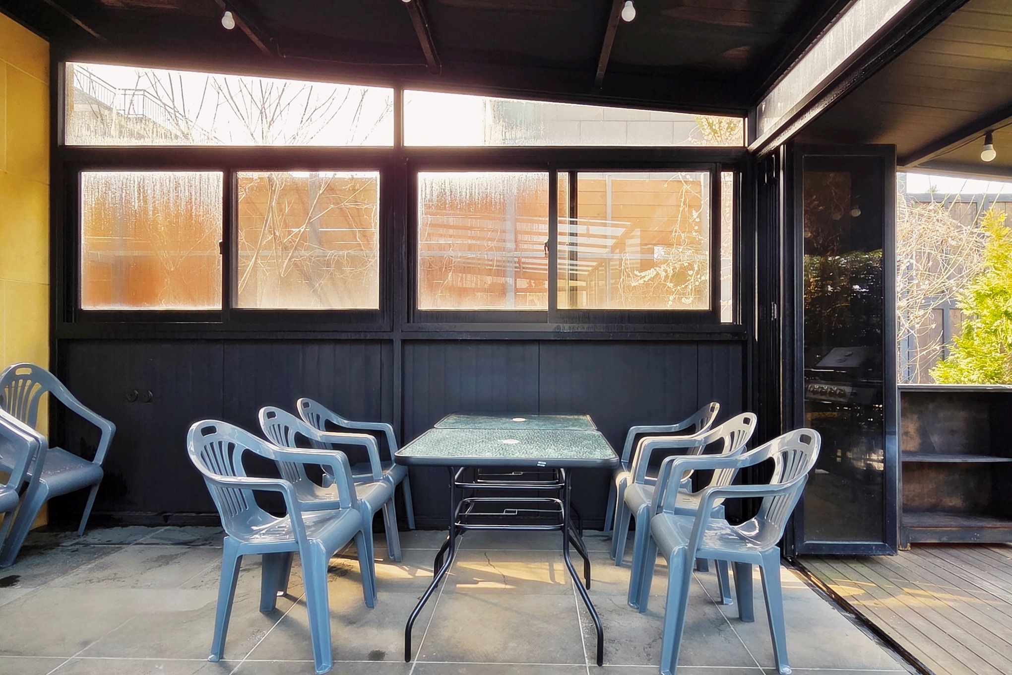 Un área de estar al aire libre con una mesa, sillas, ventanas grandes y vistas al exterior.