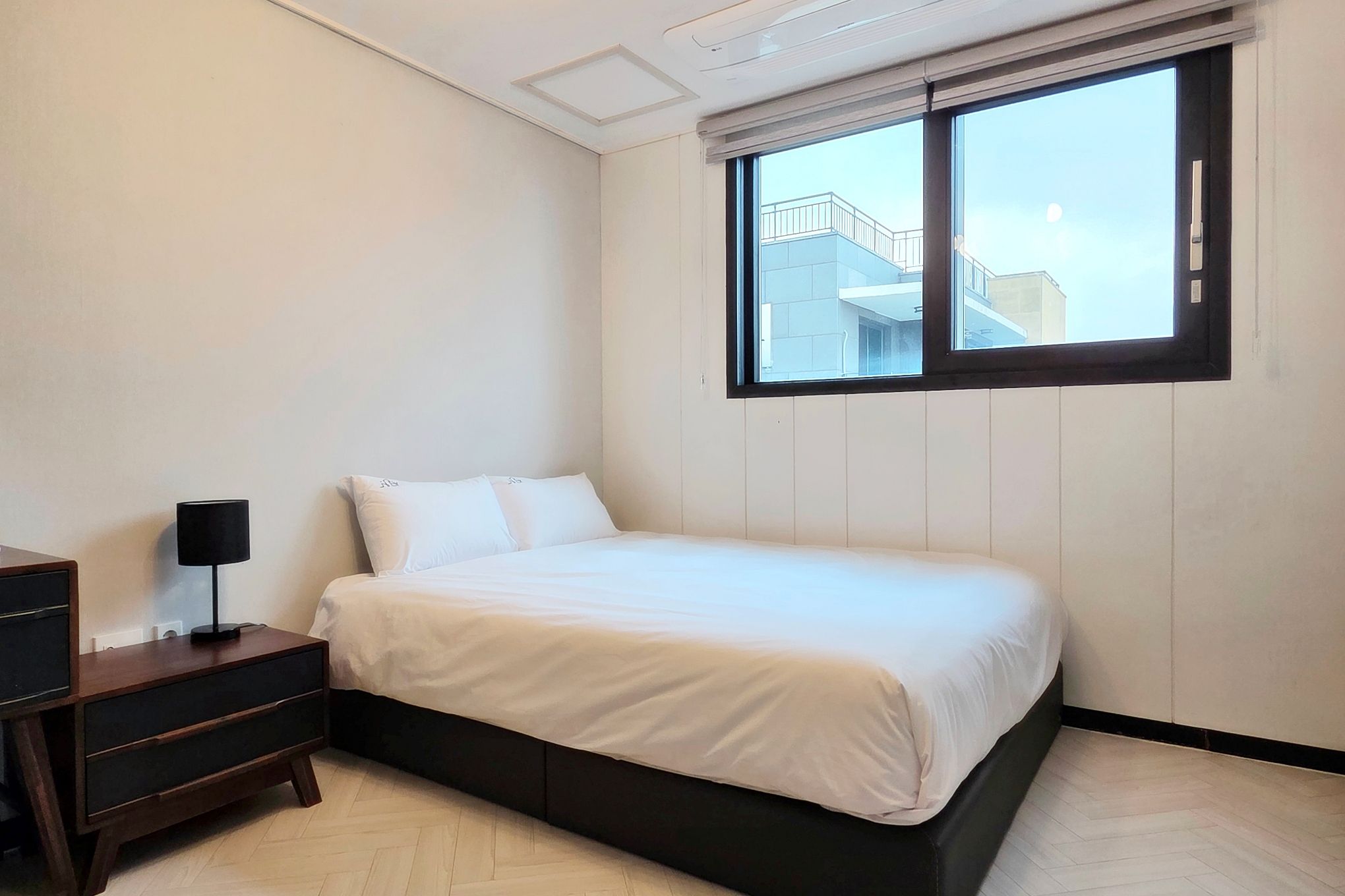 Un dormitorio con cama, mesita de noche, lámpara y ventana con vistas a edificios.