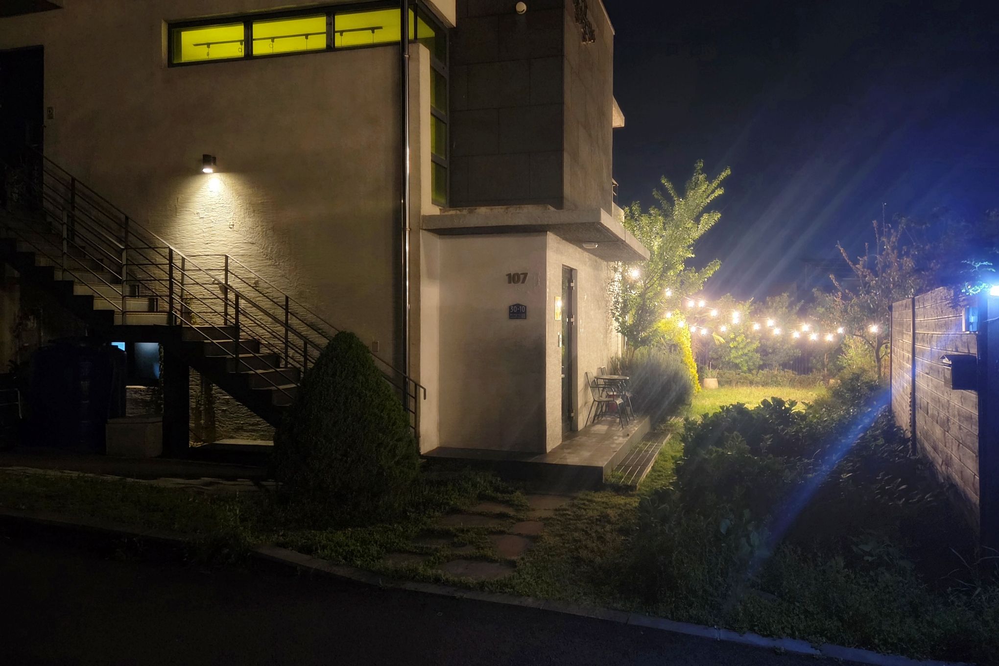Ein Gebäude mit einer Treppe, einer Tür mit der Aufschrift '107' und einem gut beleuchteten Außenbereich mit Lichterketten.