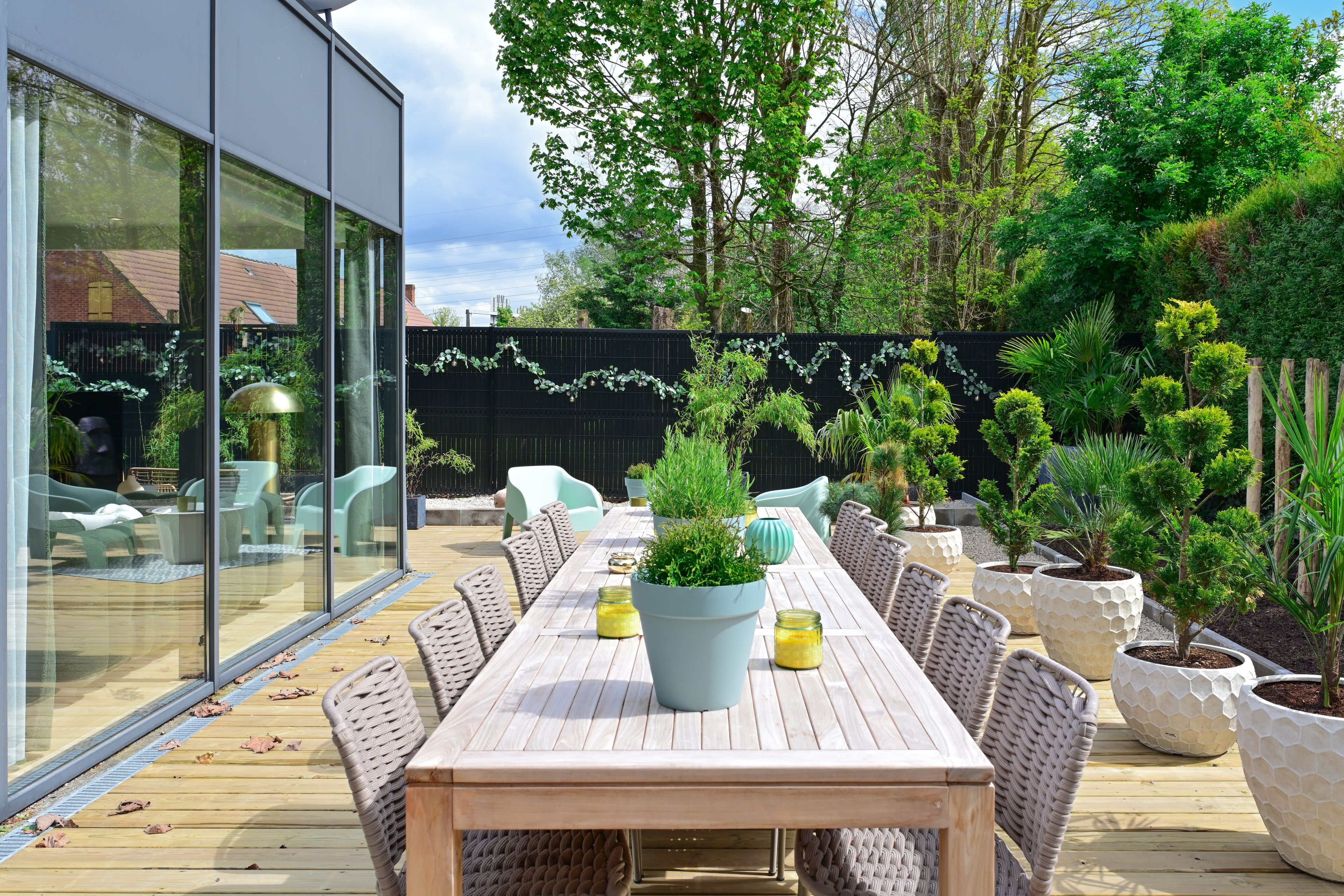 An outdoor dining area with a long wooden table and wicker chairs, surrounded by potted plants and a glass wall.