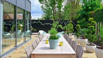 An outdoor dining area with a long wooden table and wicker chairs, surrounded by potted plants and a glass wall.