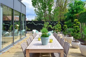 An outdoor dining area with a long wooden table and wicker chairs, surrounded by potted plants and a glass wall. - Extraordinaire Villa 5 étoiles 12 personnes 7 min Lille (Marcq-en-Barœul)