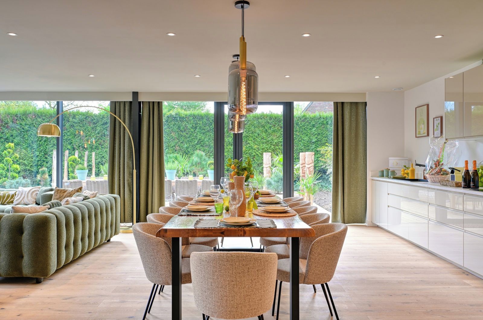 A modern dining area with a long table set for six, surrounded by chairs, with a view of greenery outside.