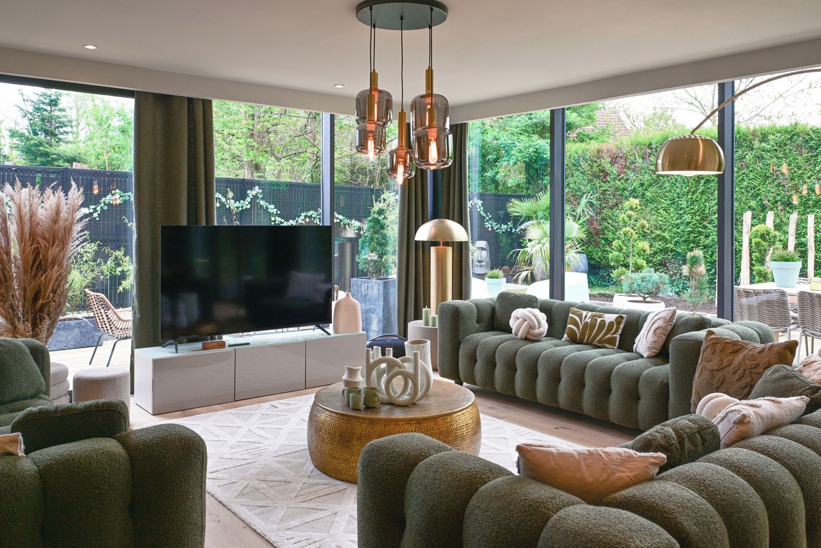 A modern living room with a large flat-screen TV, comfortable seating, and a view of a lush garden through large windows.
