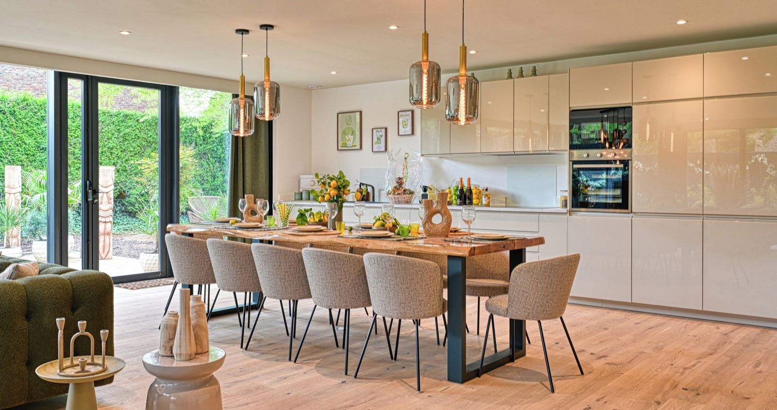 A modern kitchen with a dining area, featuring a large island, pendant lights, and a view of greenery outside.