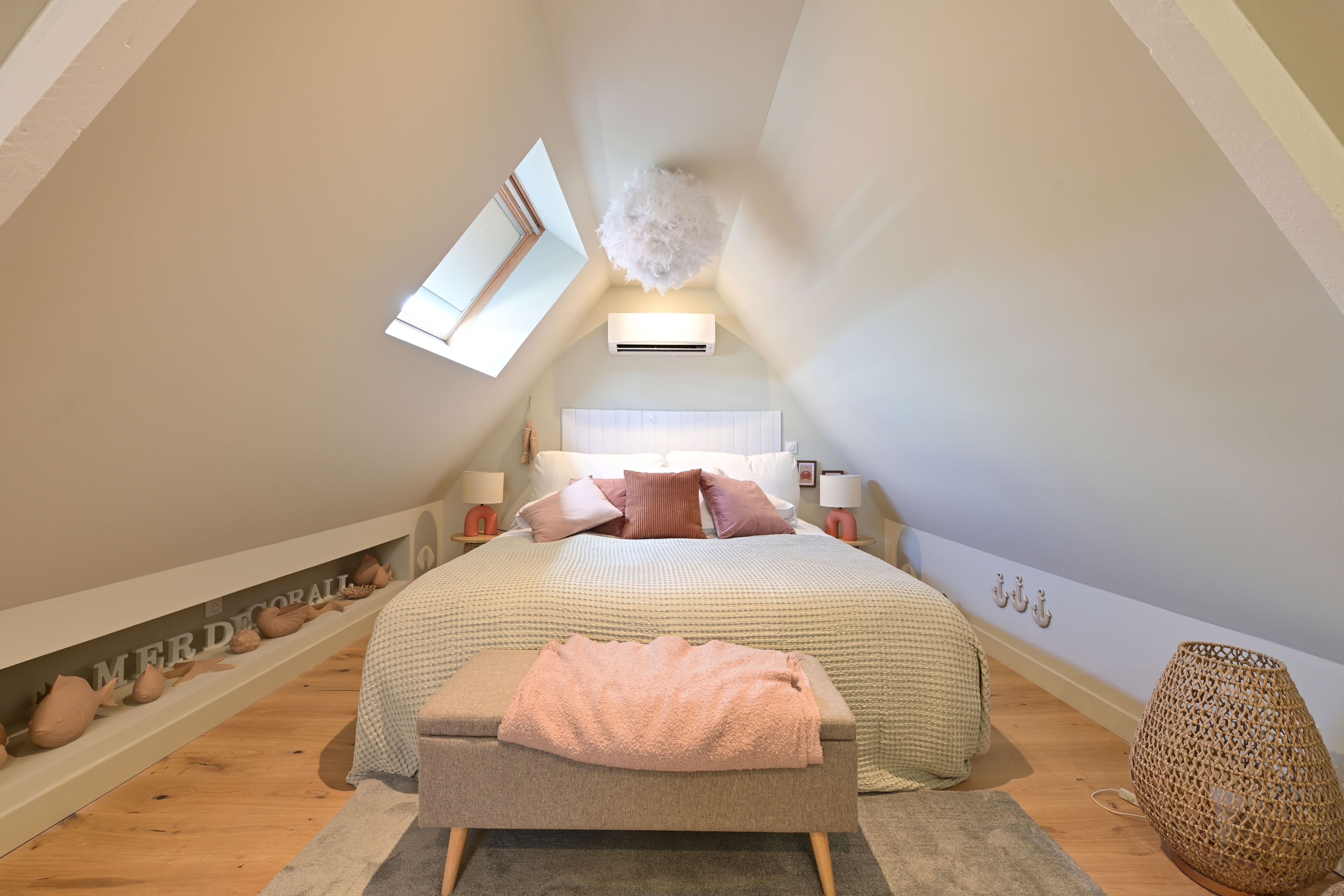 A bedroom with a sloped ceiling, a skylight, a bed with multiple pillows, a bedside table, and a decorative wall shelf.