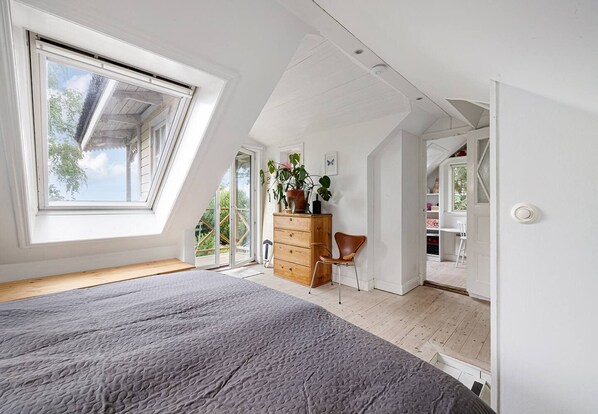 Un dormitorio con techo inclinado, un ventanal grande, una cama, un aparador y una silla.