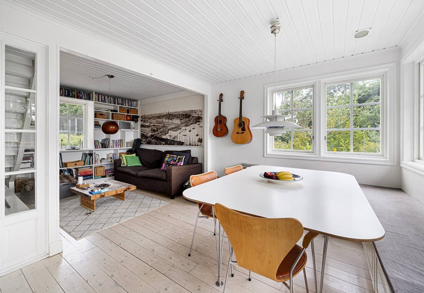 A spacious living area with a white ceiling, a dining table with chairs, a bookshelf, and a large window with a view of greenery.