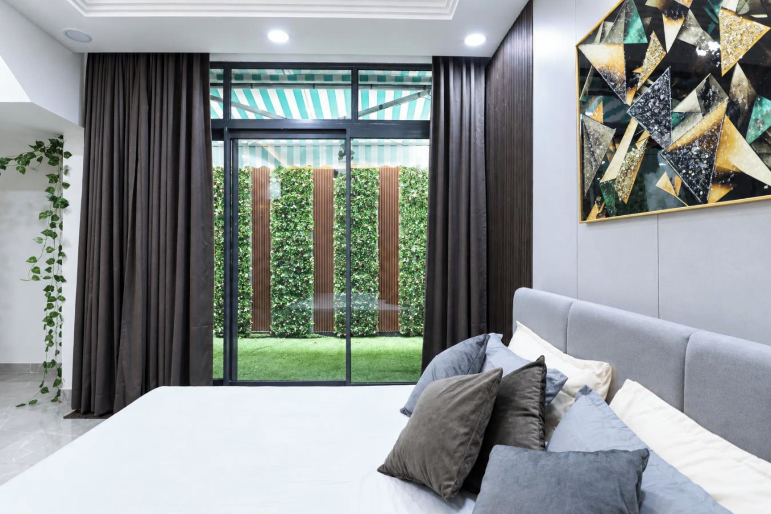 Quarto moderno com cama grande, vista para um jardim verdejante através de uma porta de vidro e uma obra de arte geométrica na parede.