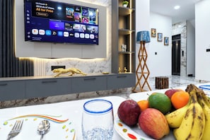 Habitación de hotel con televisor de pantalla plana mostrando diversas plataformas de streaming, suelo de baldosas de mármol, zona de comedor con una bandeja de frutas y un diseño interior moderno.