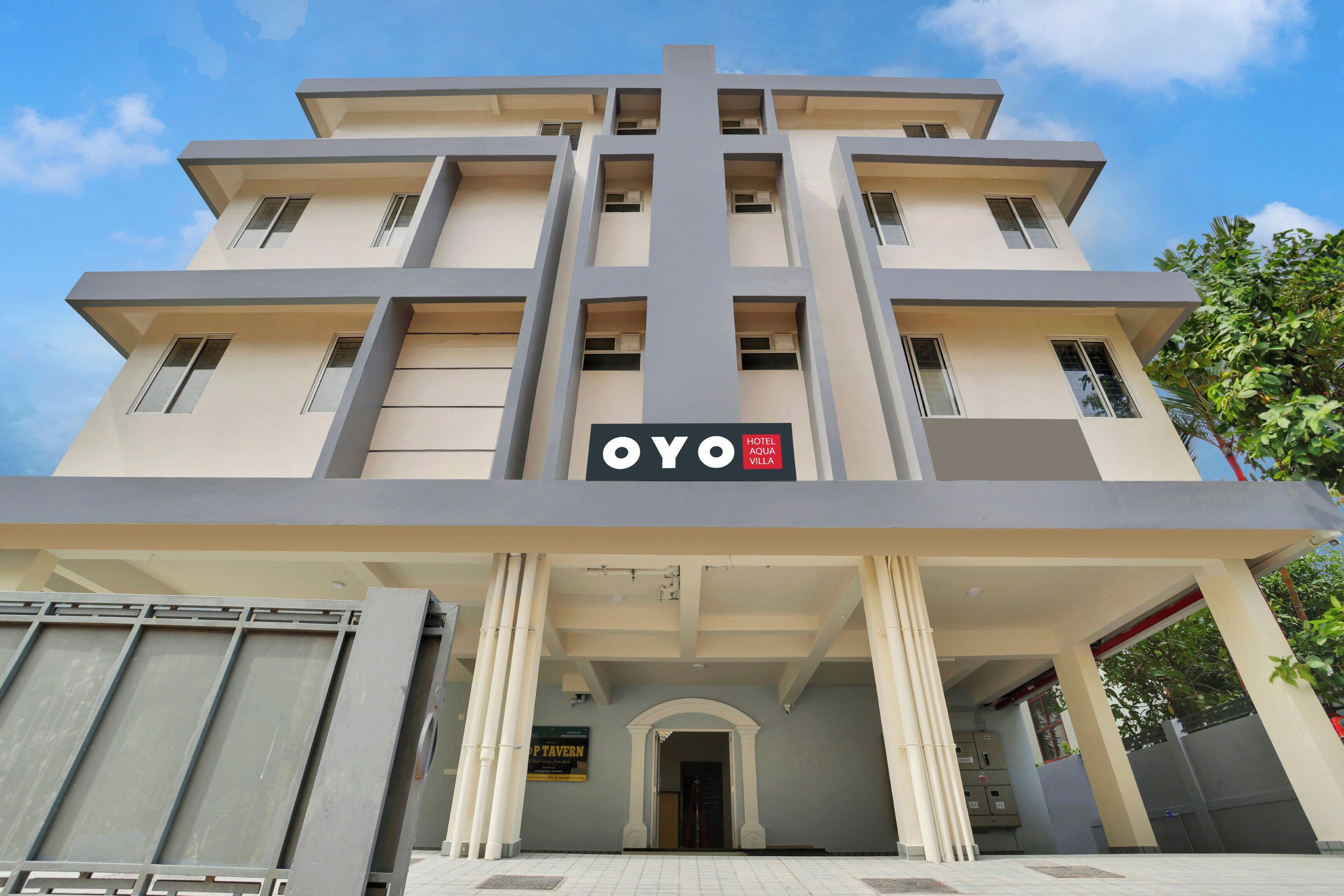 Un hotel con una fachada moderna, un letrero llamativo que dice 'OYO HOTEL AQUIA VILLA' y un diseño arquitectónico simétrico con columnas en la entrada.