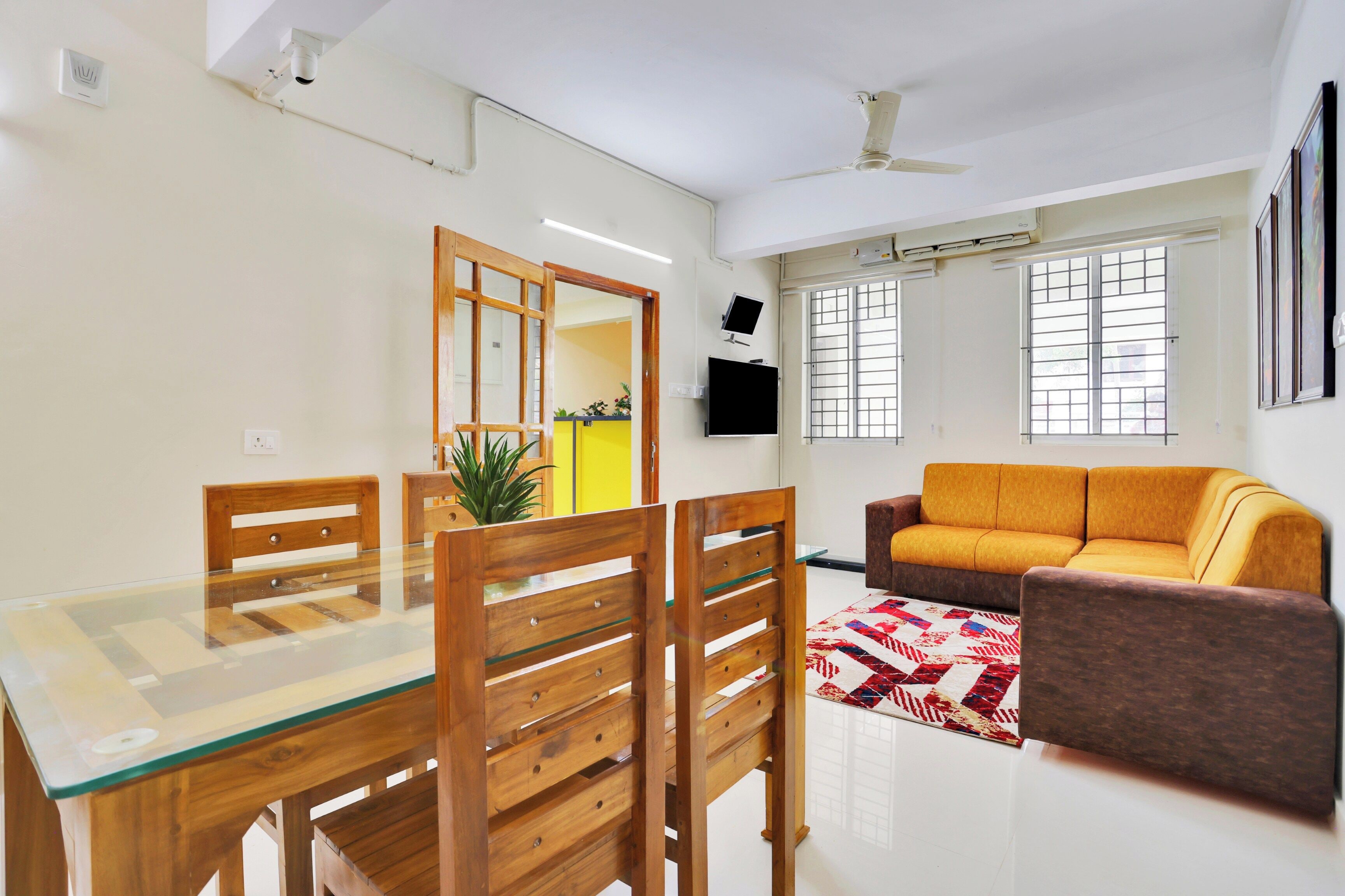 A modern living room with a glass dining table, wooden chairs, a sofa, and a TV mounted on the wall.