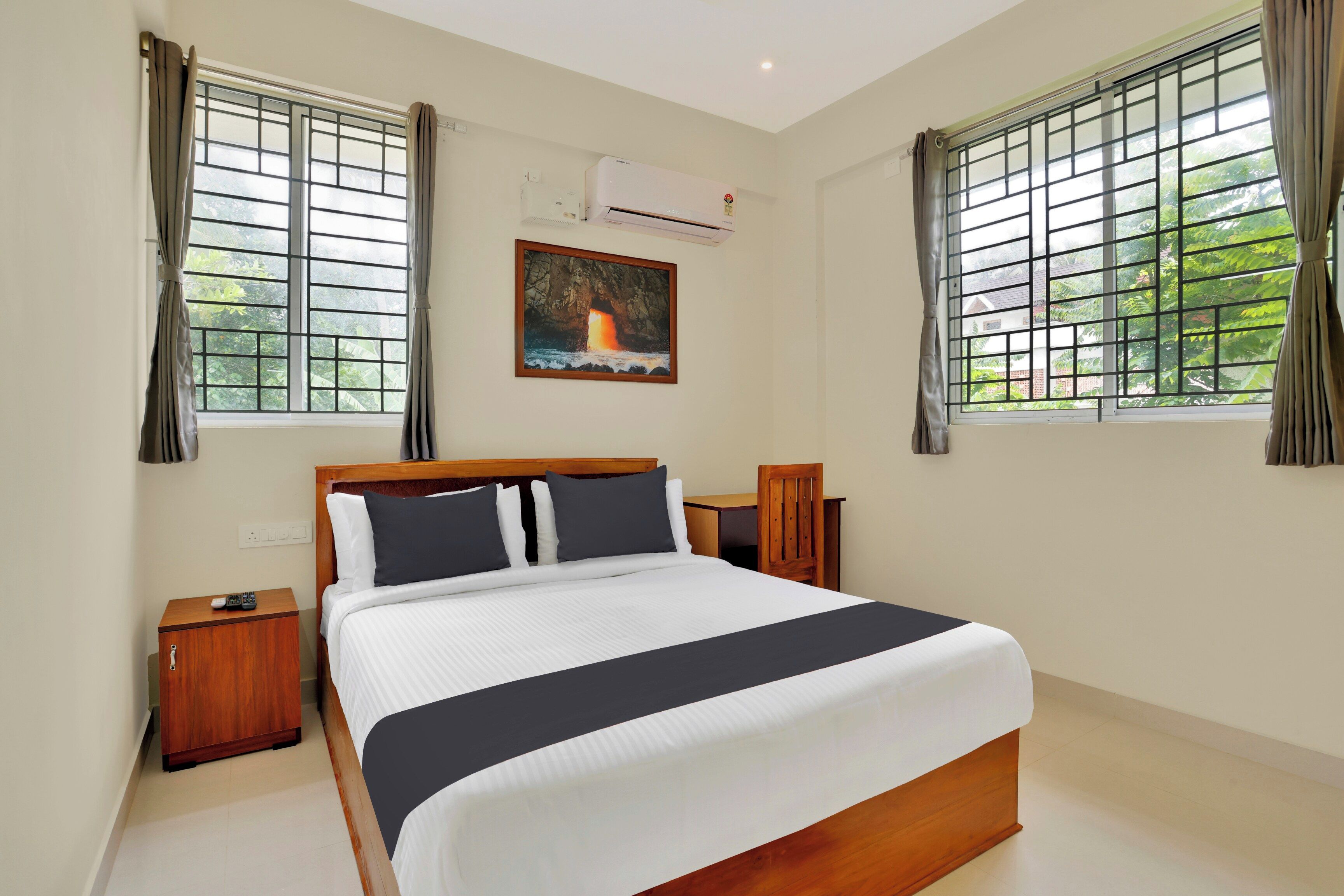 A hotel room with a bed, bedside tables, a painting, and windows with curtains.