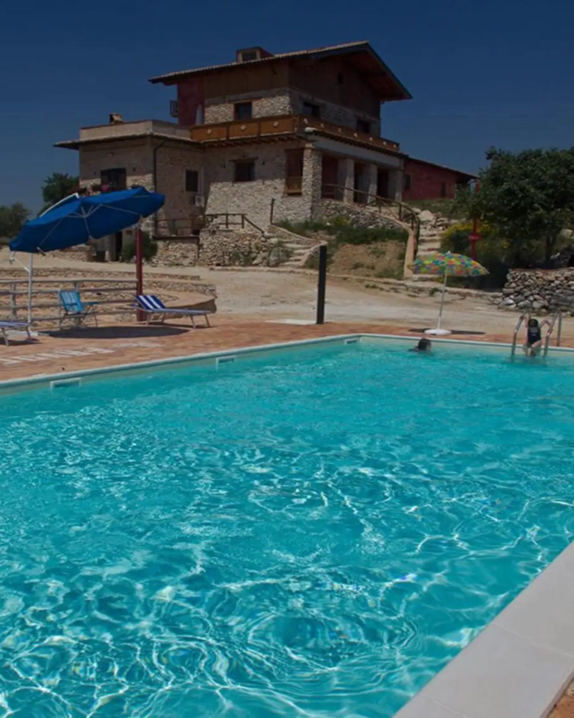 A swimming pool with lounge chairs and umbrellas, a stone building in the background, and a clear sky.