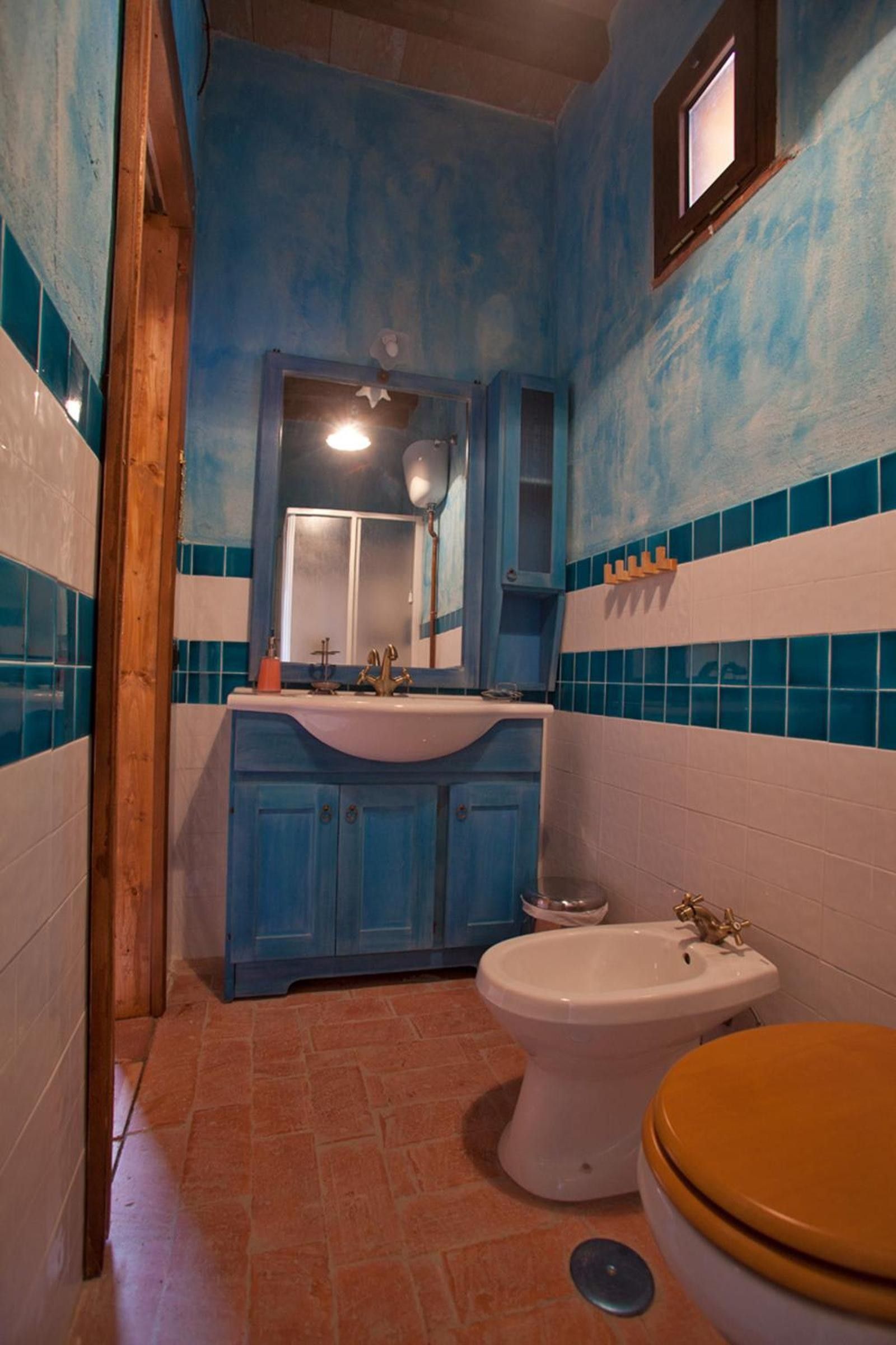 Un baño con pared de azulejos azules y blancos, una puerta de madera, un lavamanos blanco, un inodoro blanco y un ventanal pequeño.