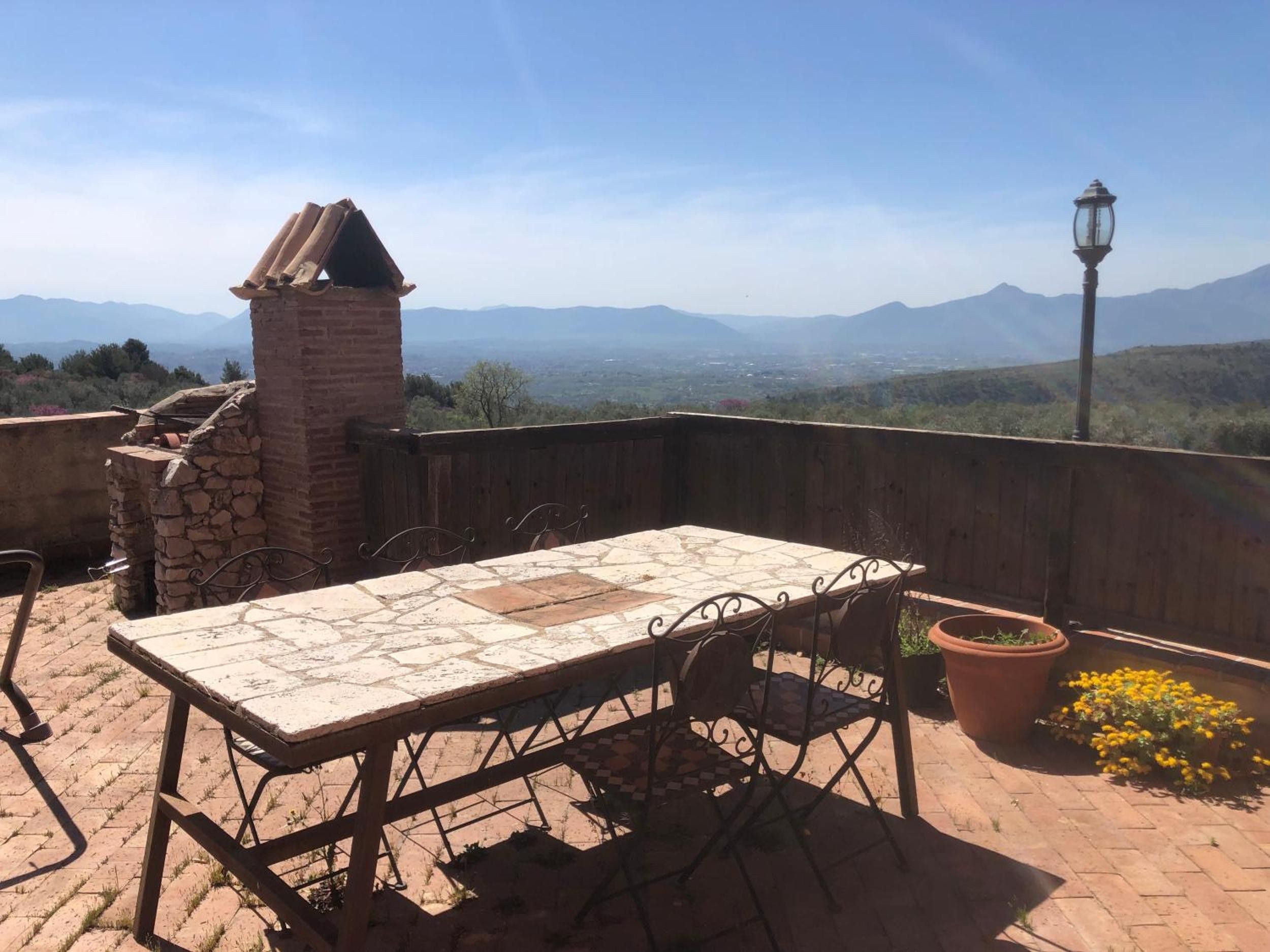 Un patio al aire libre con una mesa y sillas, un horno de ladrillo y vistas a montañas distantes.