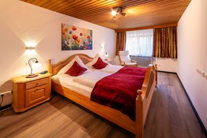 A hotel room with a wooden bed, a nightstand, a lamp, a painting, and a window with curtains. - Dahoam Holiday Apartment at Schneidersepp with a terrace, bathroom with shower and toilet, Wi-Fi, max. 3 guests (Ruhpolding)