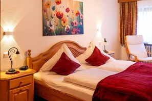Ein Hotelzimmer mit einem hölzernen Bett, zwei Nachttischlampen, einem floralen Gemälde an der Wand, einem Sessel und einem Fenster mit Vorhängen.