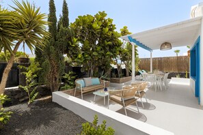 En udendørs terrasse med hvide og blå farver, med et siddeområde med sofa og stole, et lille bord og en pergola med hængende lys.