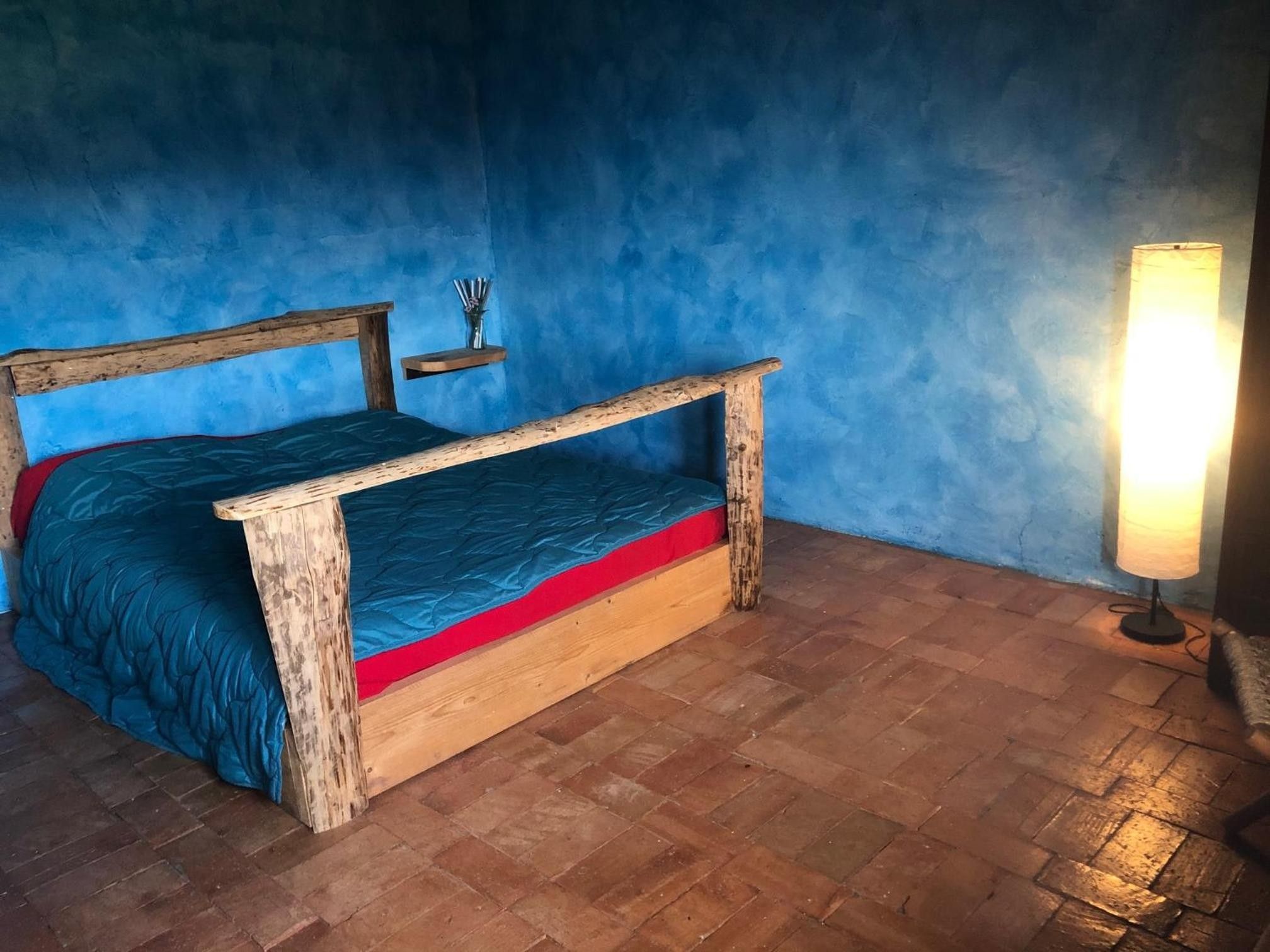 Ein Bett mit Holzrahmen und blauer Matratze in einem Zimmer mit blauen Wänden und einem Ziegelboden.