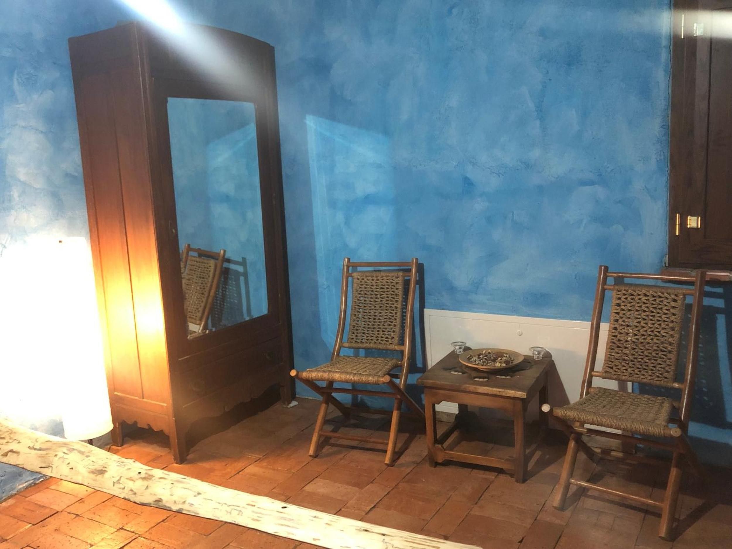 A room with a blue wall, two wicker chairs, a small table with a bowl and a candle, and a tall cabinet.