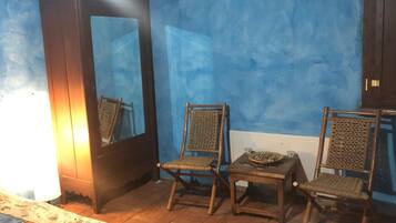 A room with a blue wall, two wicker chairs, a small table with a bowl and a candle, and a tall cabinet.